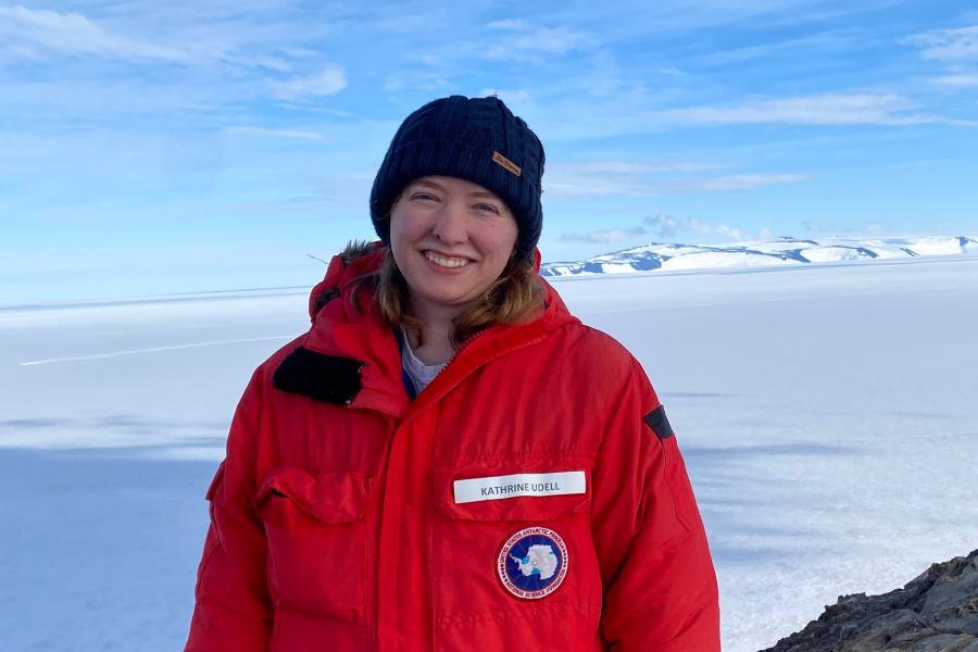 A photo of Kathrine Udell Lopez posing in a red coat in Antarctica with a snow-covered landscape in the background