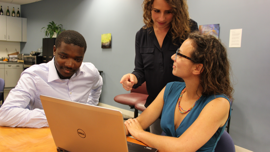 A man and a woman sitting at a table with another women standing with all three looking at a laptop screen and talking