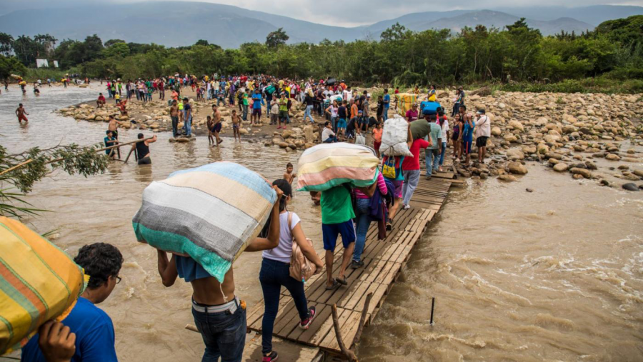 People crossing a makeshift footbridge over a flooded area, carrying their belongings