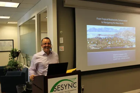 Roberto Delgado standing behind a podium in front of a presentation projected on a screen