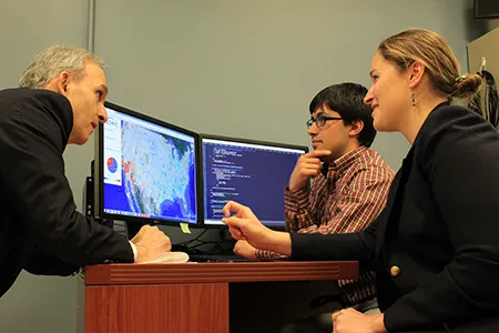 Two man and a woman sitting around a desk with a computer with two monitors